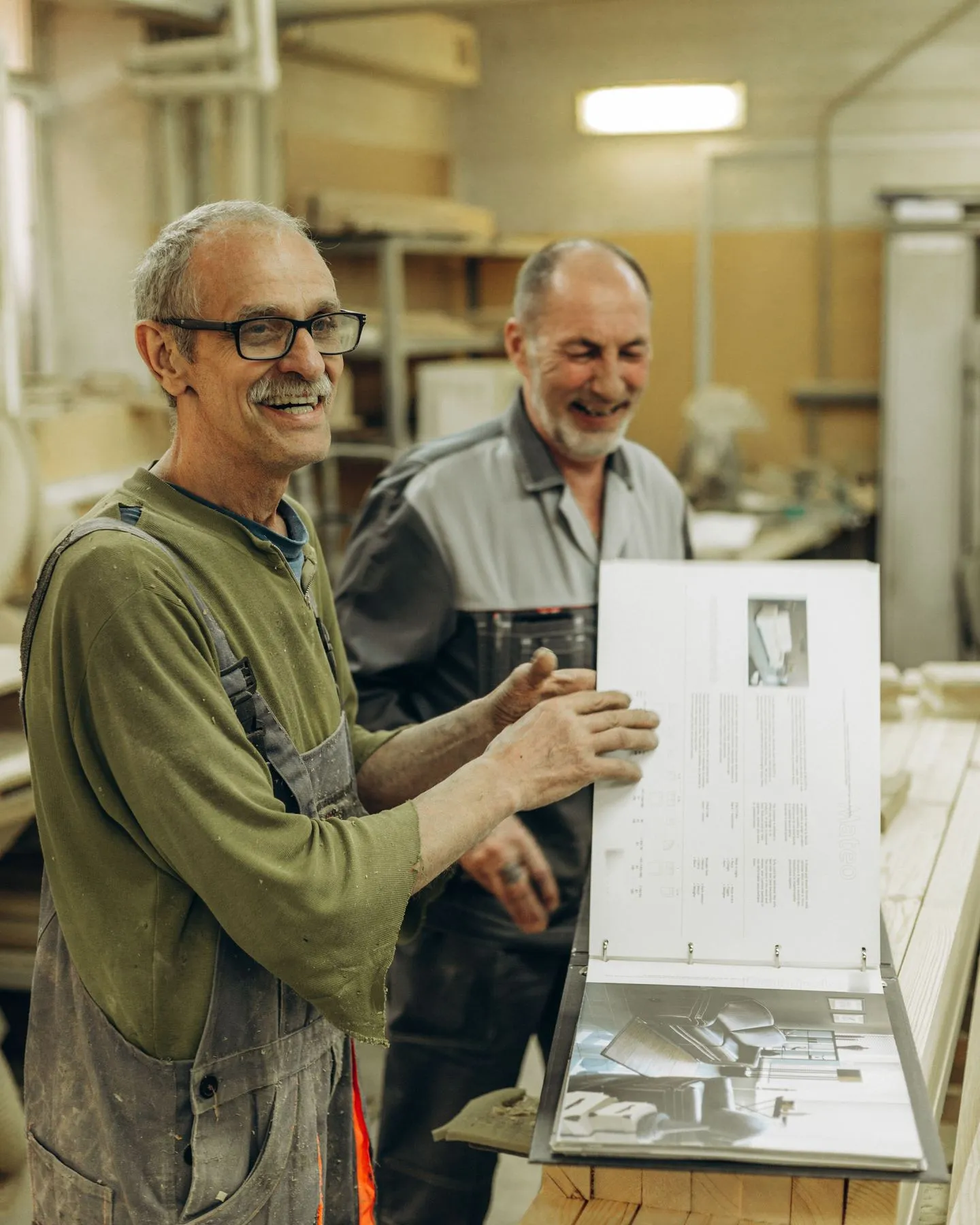 Two men in work clothes laughing as they page through a binder of product specs in a woodworking shop